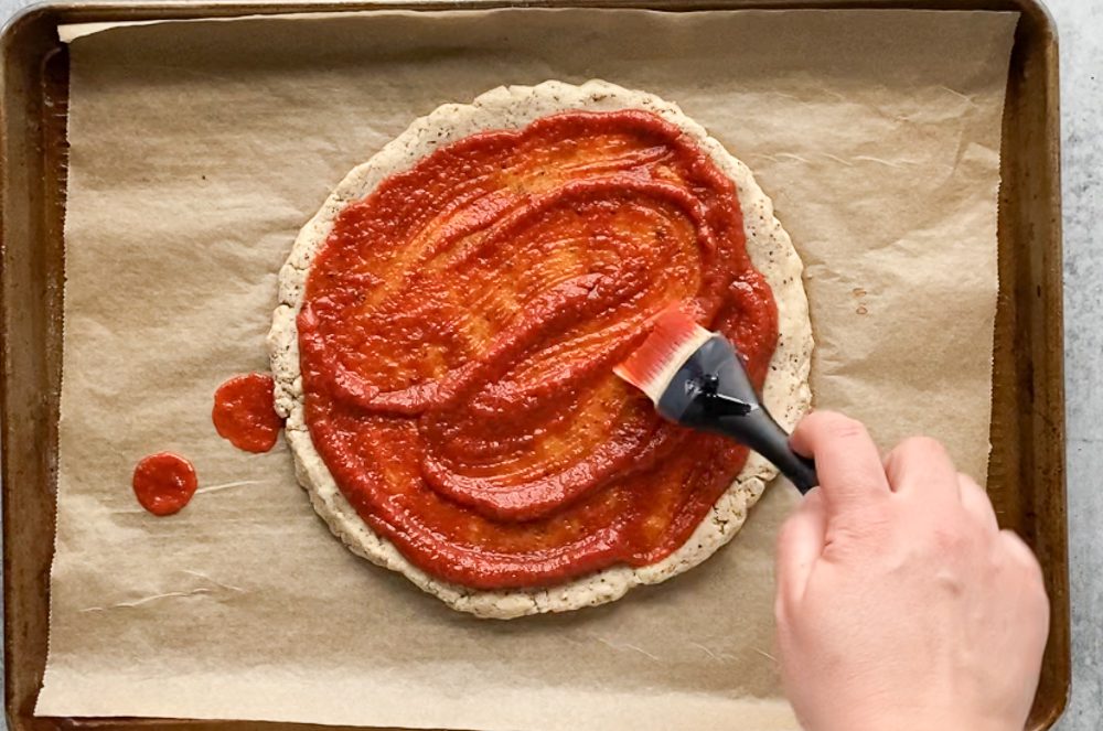 hand brushing tomato sauce on pizza crust on parchment lined baking sheet