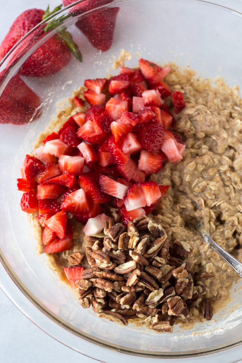 banana bread batter in glass bowl with chopped pecans and chopped strawberries with a spoon and whole strawberries on the side