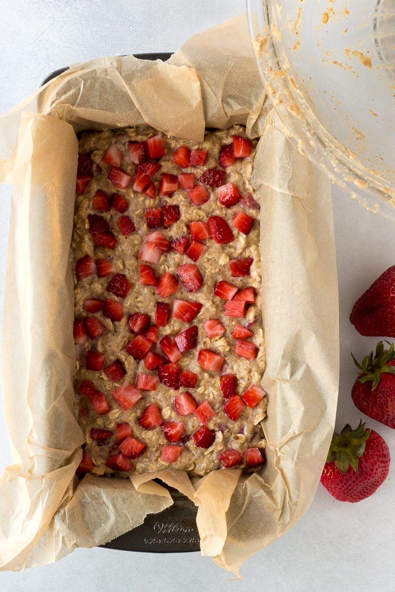 Strawberry Banana Bread batter spread into parchment lined bread pan with mixing bowl and whole strawberries on the side