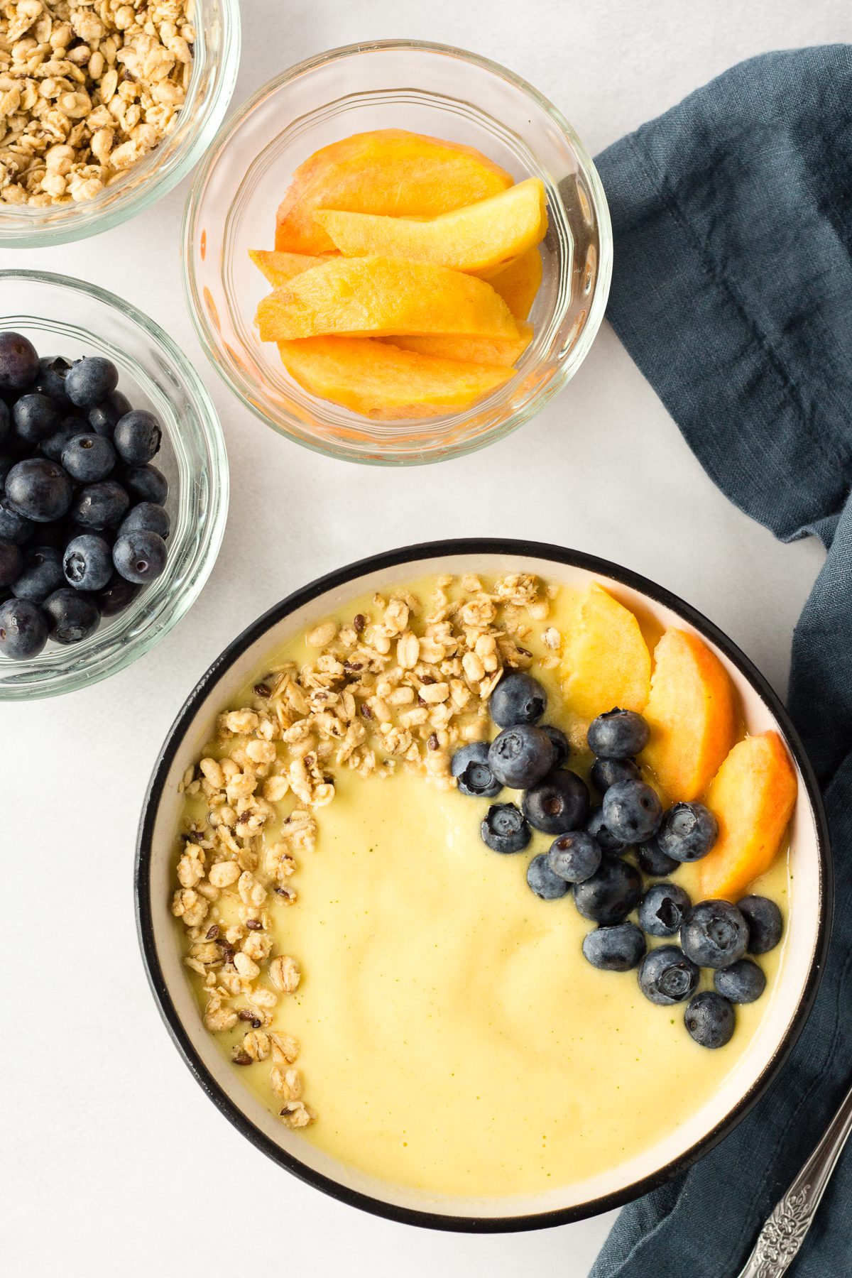 Peach Mango Smoothie Bowl served in bowl topped with blueberries, sliced peaches, and granola with spoon and blue towel 