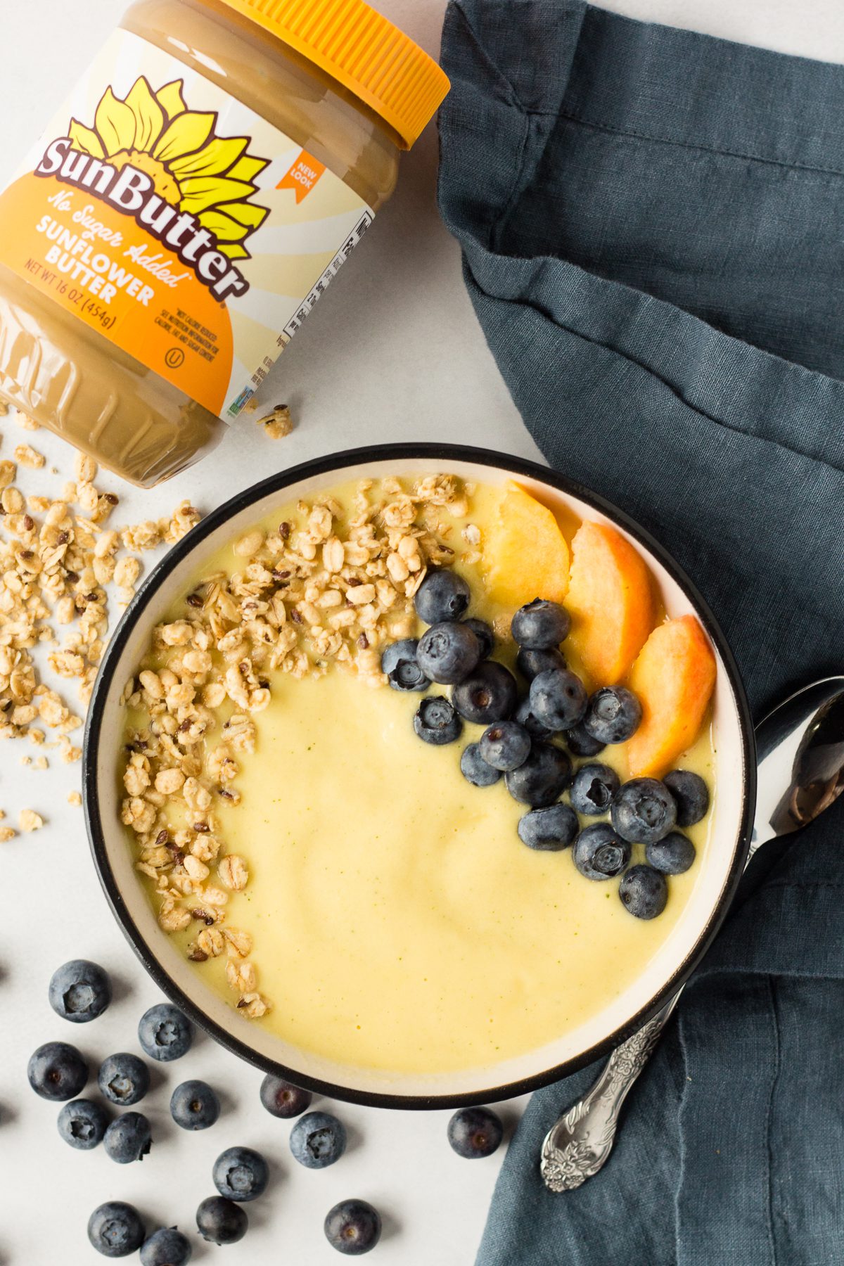 Peach Mango Smoothie Bowl served in bowl topped with blueberries, sliced peaches, and granola with spoon and blue towel 