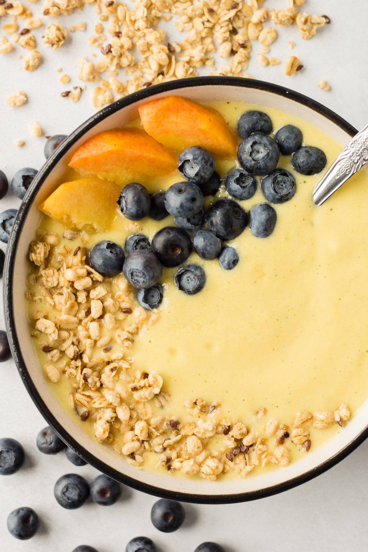 Peach Mango Smoothie Bowl served in bowl topped with blueberries, sliced peaches, and granola with spoon and blue towel 