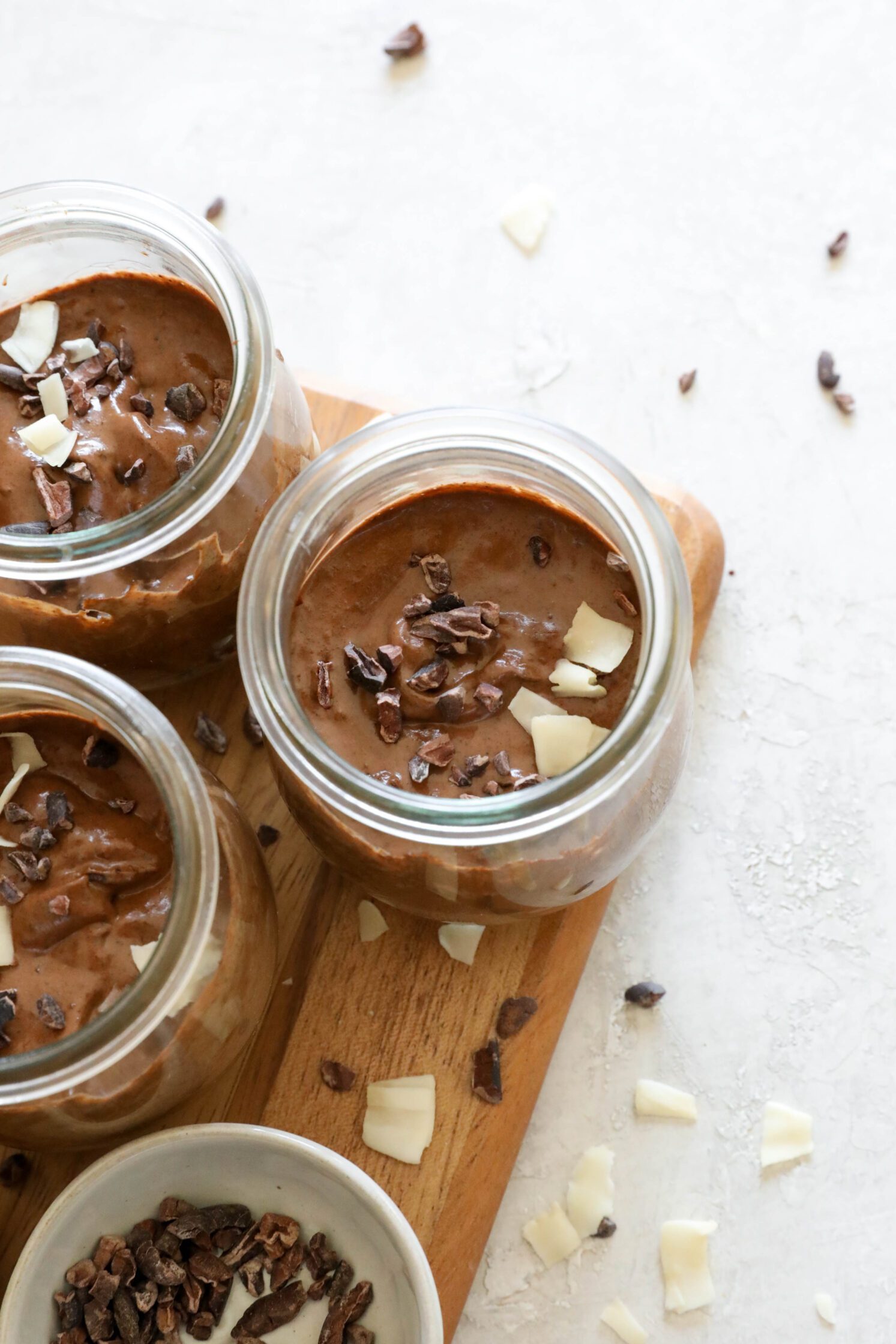 Chocolate Chia Pudding in mason jars on wooden board with bowl of cacao nibs, bowl of chia seeds, and scattered cacao nibs and toasted coconut with two spoons on the side