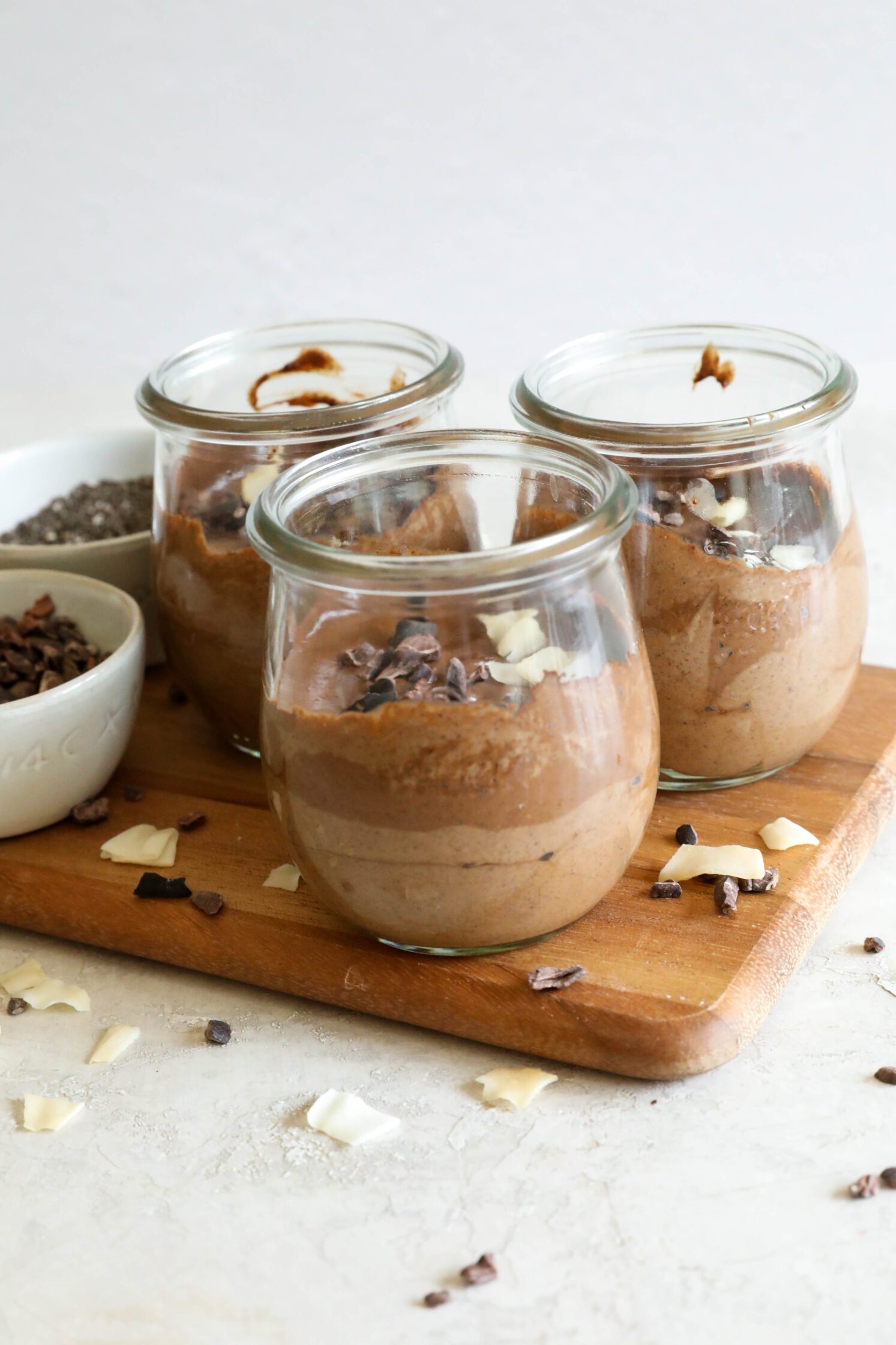 side on chocolate chia pudding in mason jars on wooden board with bowl of cacao nibs, bowl of chia seeds, and scattered cacao nibs and toasted coconut