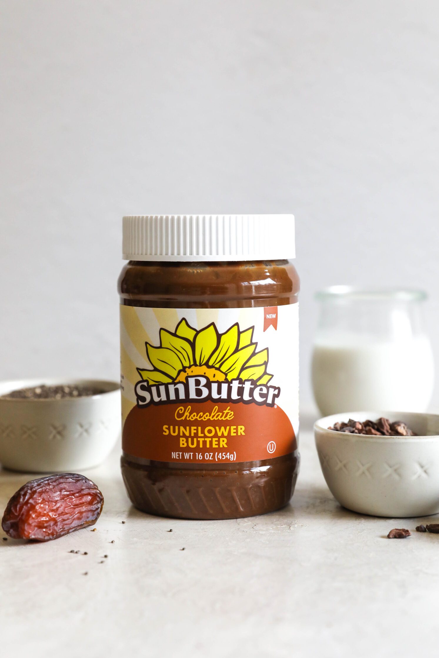 Chocolate SunButter jar on board with cacao nibs and chia seeds and Medool date and glass of almond milk