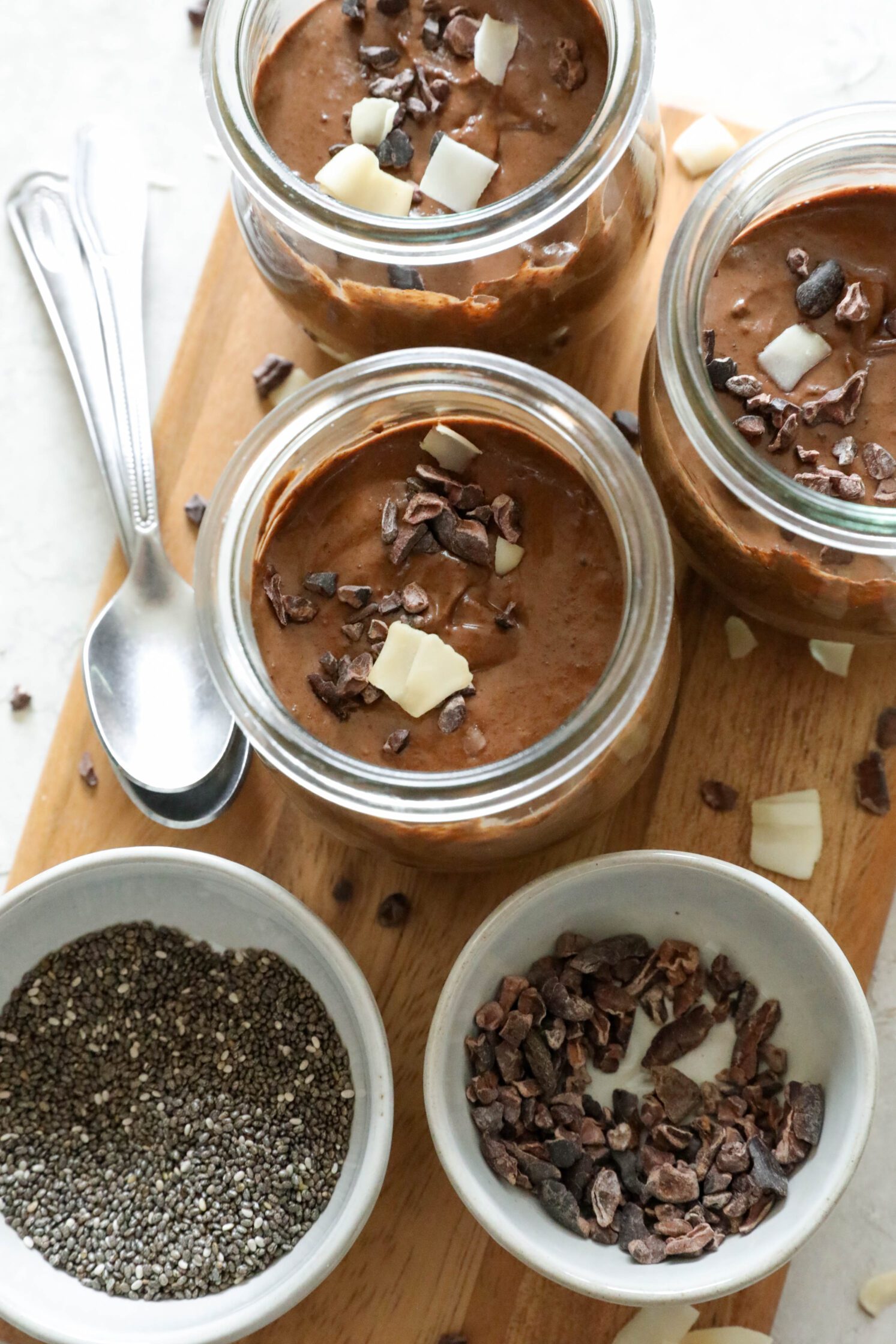 Blended Cacao Chia Pudding in mason jars on wooden board with bowl of cacao nibs, bowl of chia seeds, and scattered cacao nibs and toasted coconut with two spoons on the side