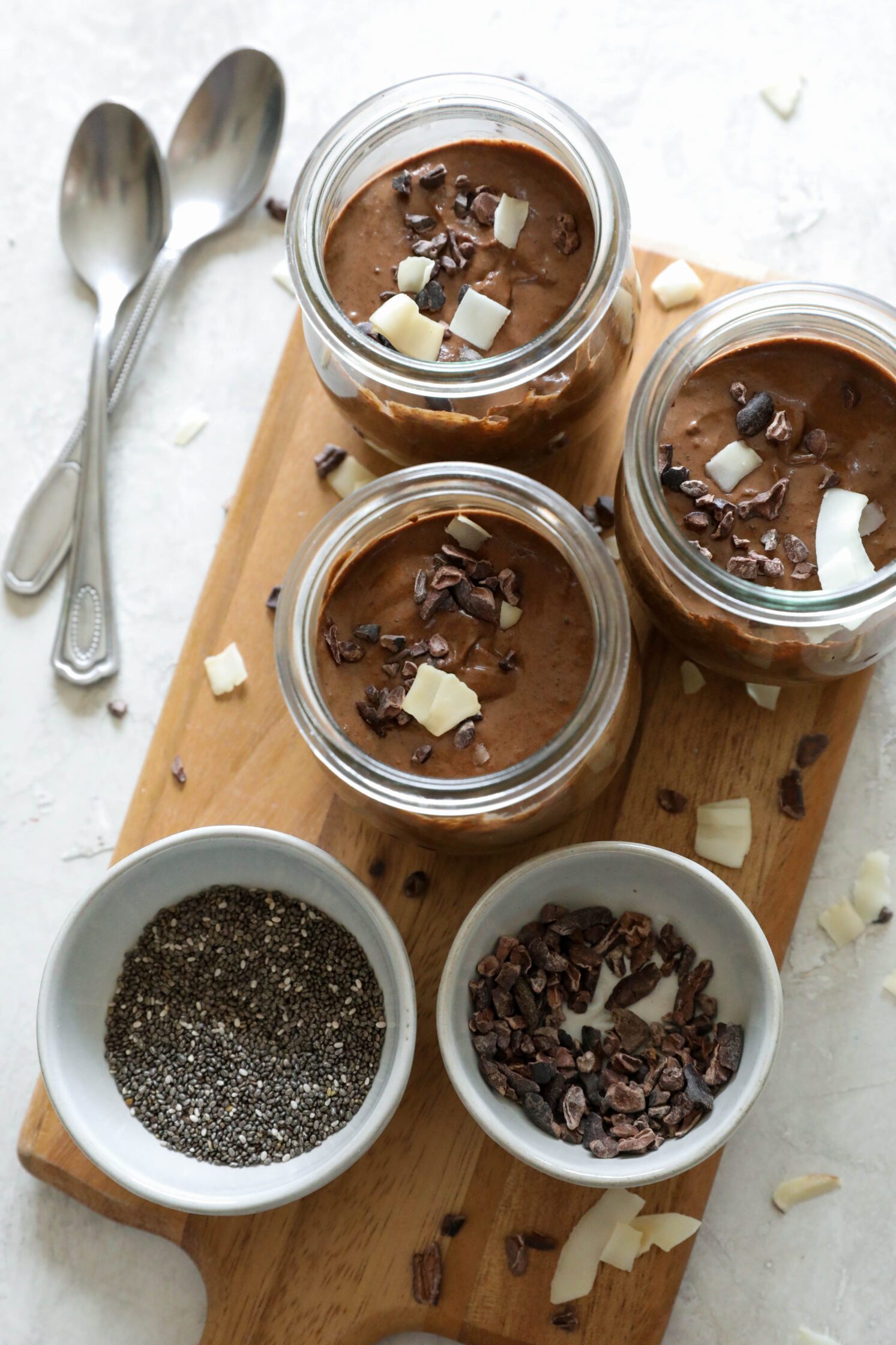 Chocolate Chia Pudding in mason jars on wooden board with bowl of cacao nibs, bowl of chia seeds, and scattered cacao nibs and toasted coconut with two spoons on the side