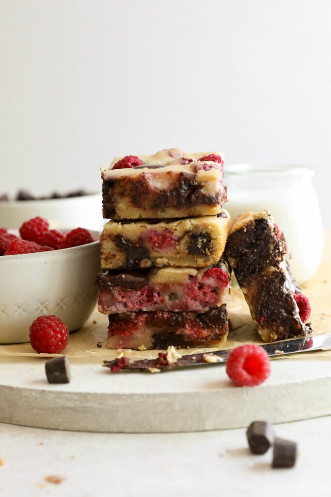 Dark Chocolate Raspberry Blondies stacked on parchment paper with knife and scattered raspberries and dark chocolate chips by Flora & Vino