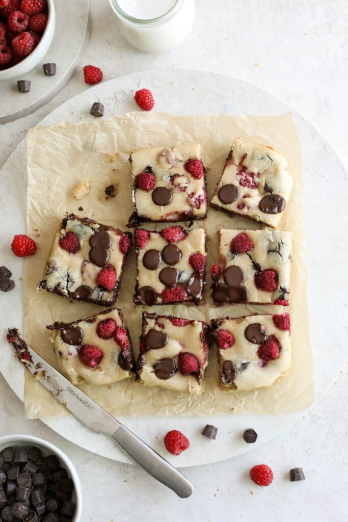 Dark Chocolate Raspberry Blondies cut into squares on parchment paper with raspberries and chocolate chips scattered by Flora & Vino