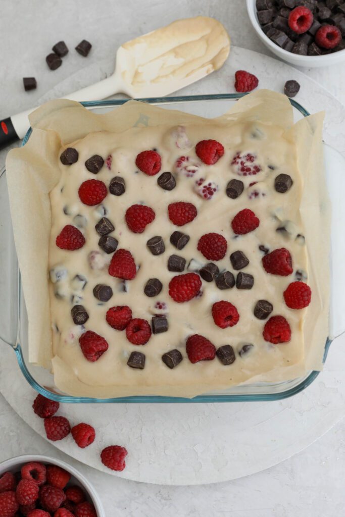 Dark Chocolate Raspberry Blondie batter in parchment lined baking dish with spatula on the side by Flora & Vino