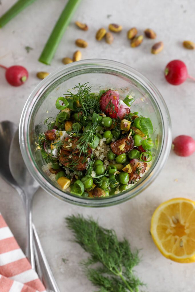 Spring Quinoa Salad Jar overhead with two spoons and dill, lemon, radishes, pistachios, and green onion with pink and white striped towel